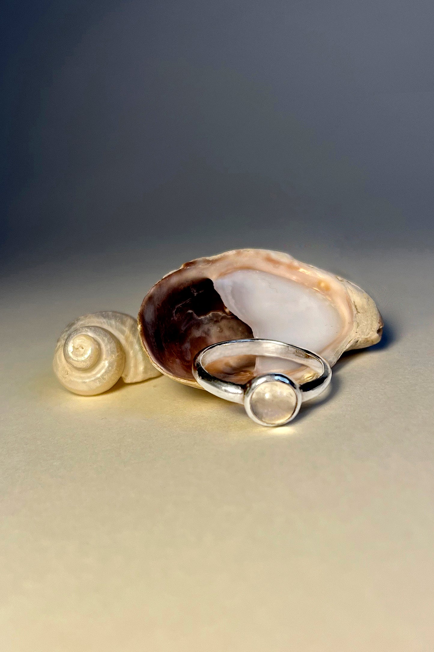 Underwater feeling photo of The Magic Mirror Moonstone Ring by CLARK has a cabochon set band with an iridescent moonstone gem resting on shell. This hand-made one-of-a-kind ring is made in Sterling Silver using the lost wax casting method. A unique gift.