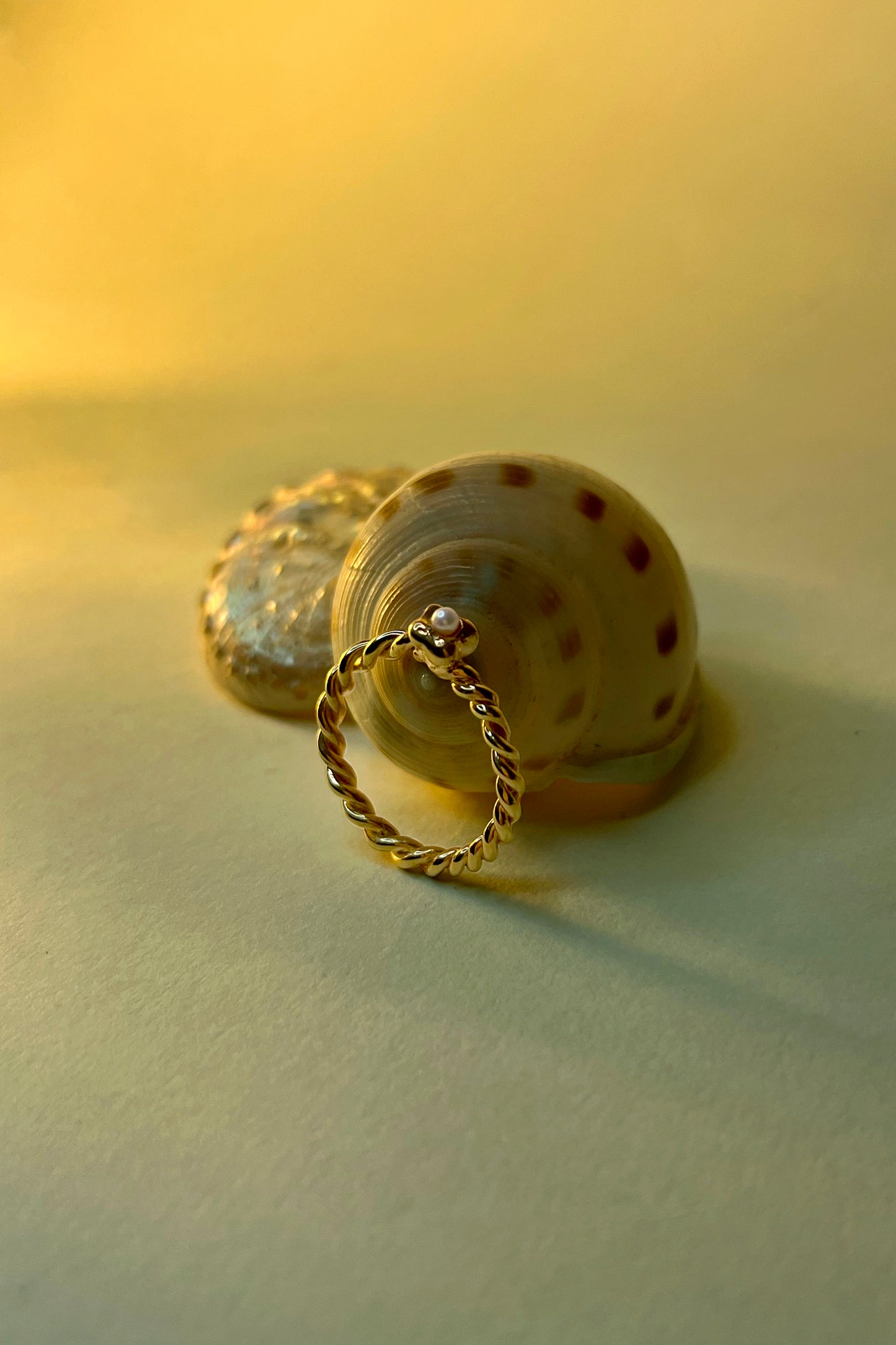 Mini Pearl Mermaid Ring by CLARK, resting against a spiral seashell. Hand craft in 18k Gold Vermeil, this delicate one-of-a-kind ring features a freshwater pearl and a twisted band, a symbolic mermaid tail.