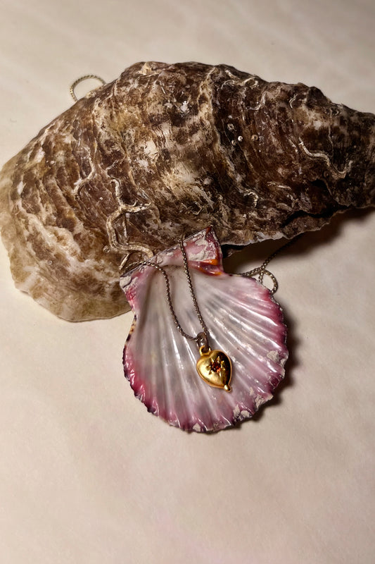 CLARK Tiny Heart necklace on a pink seashell. Handcrafted in wax and set with a garnet gemstone. Shown here in 18K Gold Vermeil