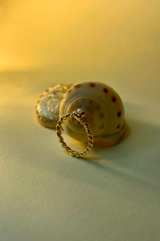 Mini Pearl Mermaid Ring by CLARK, resting against a spiral seashell. Hand craft in 18k Gold Vermeil, this delicate one-of-a-kind ring features a freshwater pearl and a twisted band, a symbolic mermaid tail. 