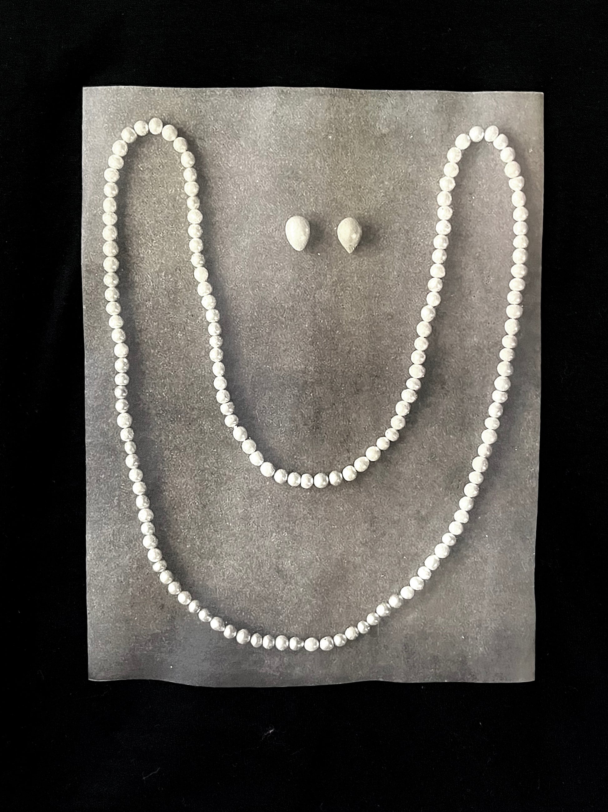 Close up of whimsical design - a pearl necklace and two pearls laid on gray pavement in the shape of a smiling face. 