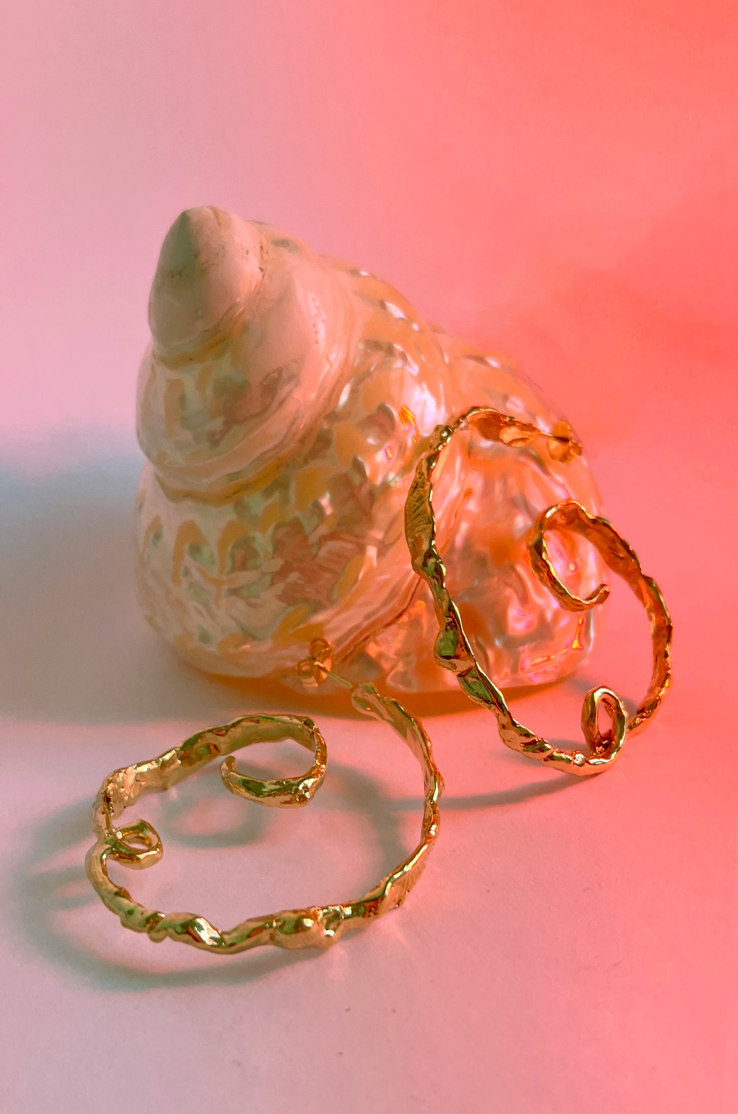 Product photo of the CLARK Andromeda Earring in 18K Gold Vermeil. Two organic hoops are balanced on a seashell. This is a red hue over this underwater scene. These earrings are hand craft using the lost wax casting technique. 