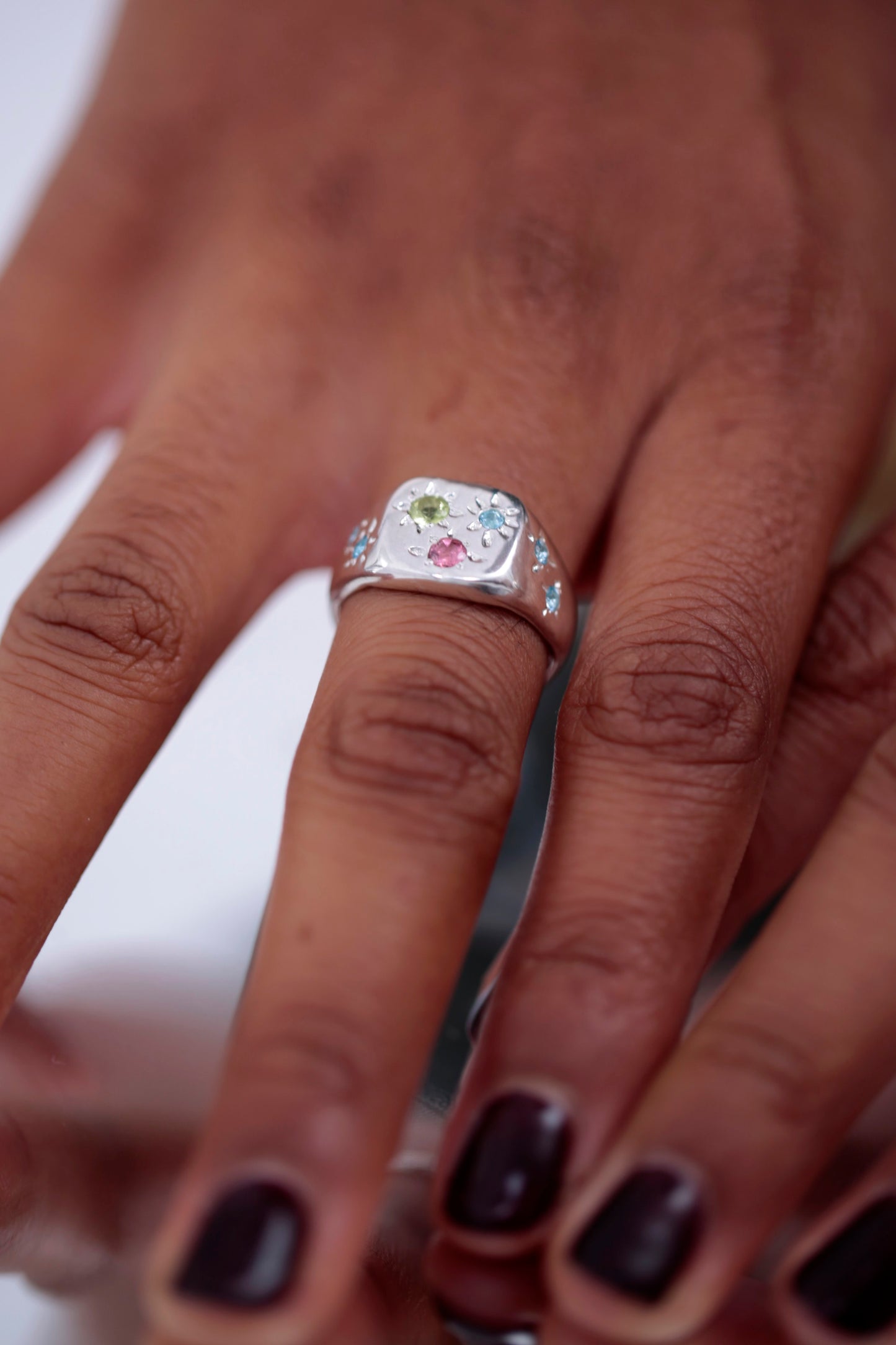 Detail image. A model's hand is placed on top of a mirrored surface. She wears the CLARK Constellation Signet ring in Sterling Silver. The colorful gems are a contrast against the luster of the Silver metal. This is a unique gift for someone with a classic and renaissance-inspired aesthetic.