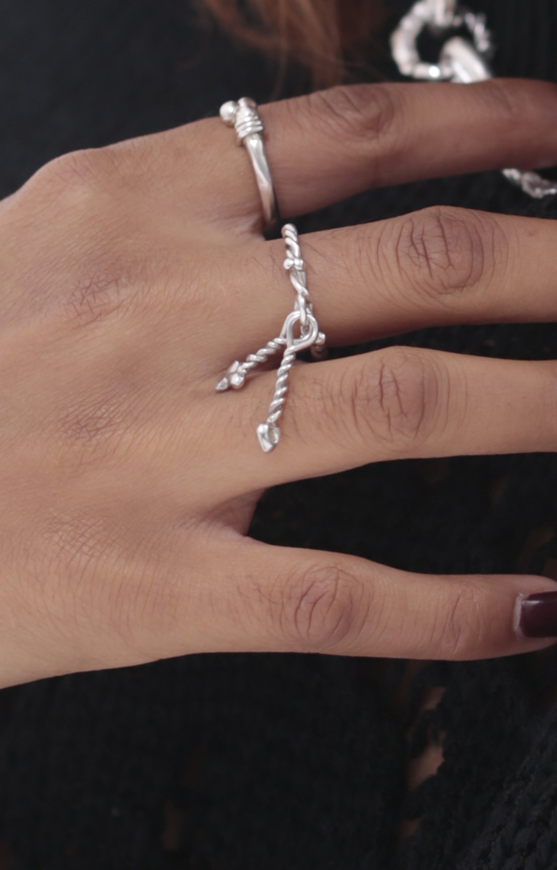 Model wears the Twisted Charm Ring by CLARK in Sterling Silver on her middle finger. Handcrafted using the lost wax casting method, this classic and simple ring shows off two dangling charms. Model also wears Eros Ring by CLARK.